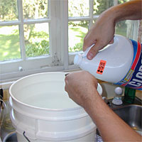Person pouring liquid from a bleach bottle into a white bucket near a window.