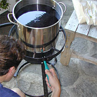 Person crouching and lighting a burner under a large pot of water on an outdoor stove.