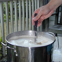 Hand stirring a metal pot of boiling liquid on a stove, with a wooden fence in the background.