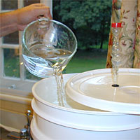 A hand pours water from a measuring cup into a white plastic barrel with a lid near a window.