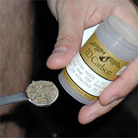 A hand holding a container labeled "ID Carlson" and filling a measuring spoon with a dry, granular substance.
