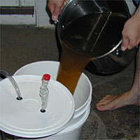 Person pouring brown liquid from a pot into a white bucket with a lid and airlock.