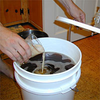 Person pouring liquid into a white fermentation bucket with a lid nearby.