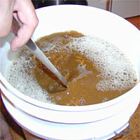 Person stirring frothy brown liquid in a white bucket with a spoon.