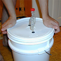 Hands holding a white plastic bucket with a fitted lid featuring an airlock for fermentation.