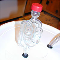 Glass fermentation airlock with a red cap on a white lid.