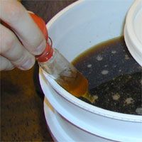Hand holding a small vial containing a amber colored liquid over a container filled with dark liquid.