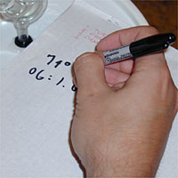 A hand writing numbers on a white paper with a black marker. A glass pipette is visible in the top left corner.