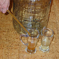 A person stirring liquid in a small glass pitcher with another clear liquid pitcher beside it, with a large jug in the background.