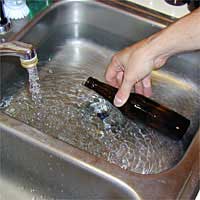 Hand rinsing a dark bottle under running water in a metal sink.