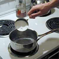 Hand pouring corn sugar powder from a measuring cup into a pot of boiling water on a stove.