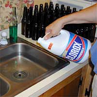 Person pouring Clorox bleach into a kitchen sink beside several empty bottles on the counter.