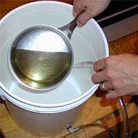 Hands holding a metal ladle scooping liquid from a white bucket.