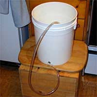 White bucket on a wooden seat with a clear hose attached at the bottom, extending downward.