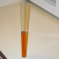Glass test tube filled with amber liquid, standing upright on a white surface.