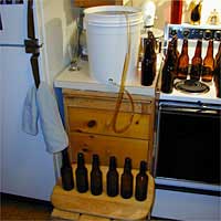 Bottling setup in a kitchen with a large bucket, brown bottles on a stove, and more bottles on a stool.