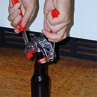 Hands using a red and silver bottle capper to seal a glass bottle on a wooden floor.