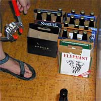 Person wearing sandals using a manual bottle capper to cap a bottle near several packs of beer on the floor.